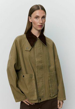 Jeune personne aux cheveux raides porte une veste oversize beige avec un col marron foncé, debout devant un fond clair uni.