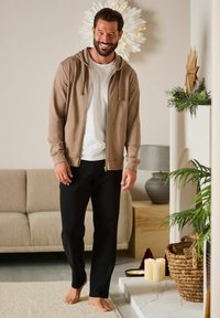 Beige lynlås hoodie over en hvid t-shirt, kombineret med sorte bukser. Bløde materialer, afslappet pasform, og en hyggelig indendørs indstilling med grønne planter.