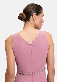 V-hals, mouwloze jurk van zachtroze stof met een decoratieve met kralen versierde tailleband en een ritsdetail op de rug, die een gladde textuur benadrukt.