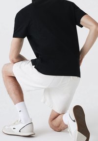 Polo noir à manches courtes, short en jean blanc, baskets blanches avec des accents gris, et chaussettes blanches. Le modèle est en position à genoux.