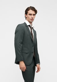 Σκούρο πράσινο κοστούμι με κομψή εφαρμογή, δύο κουμπιά και πέτο. Συνοδεύεται από λευκό πουκάμισο και γραβάτα με σχέδιο. Λείο υλικό.