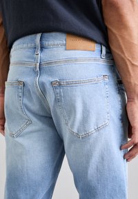 Svijetloplave traperice s kožnom etiketom na pojasu. Imaju pet džepova i vidljive šavove, uz uski kroj.