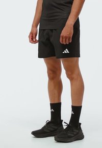 Muškarac u crnim Adidas šorcama, crnim čarapama s logom Adidas i crnim Adidas tenisicama stoji na jednobojnoj pozadini.