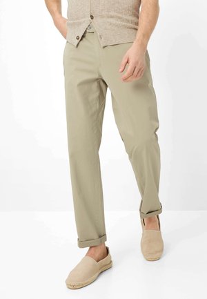 Mann trägt beige Chinos mit Aufschlägen, eine hellbraune, geknöpfte Strickjacke und beige Espadrilles zum Hineinschlüpfen vor weißem Hintergrund.