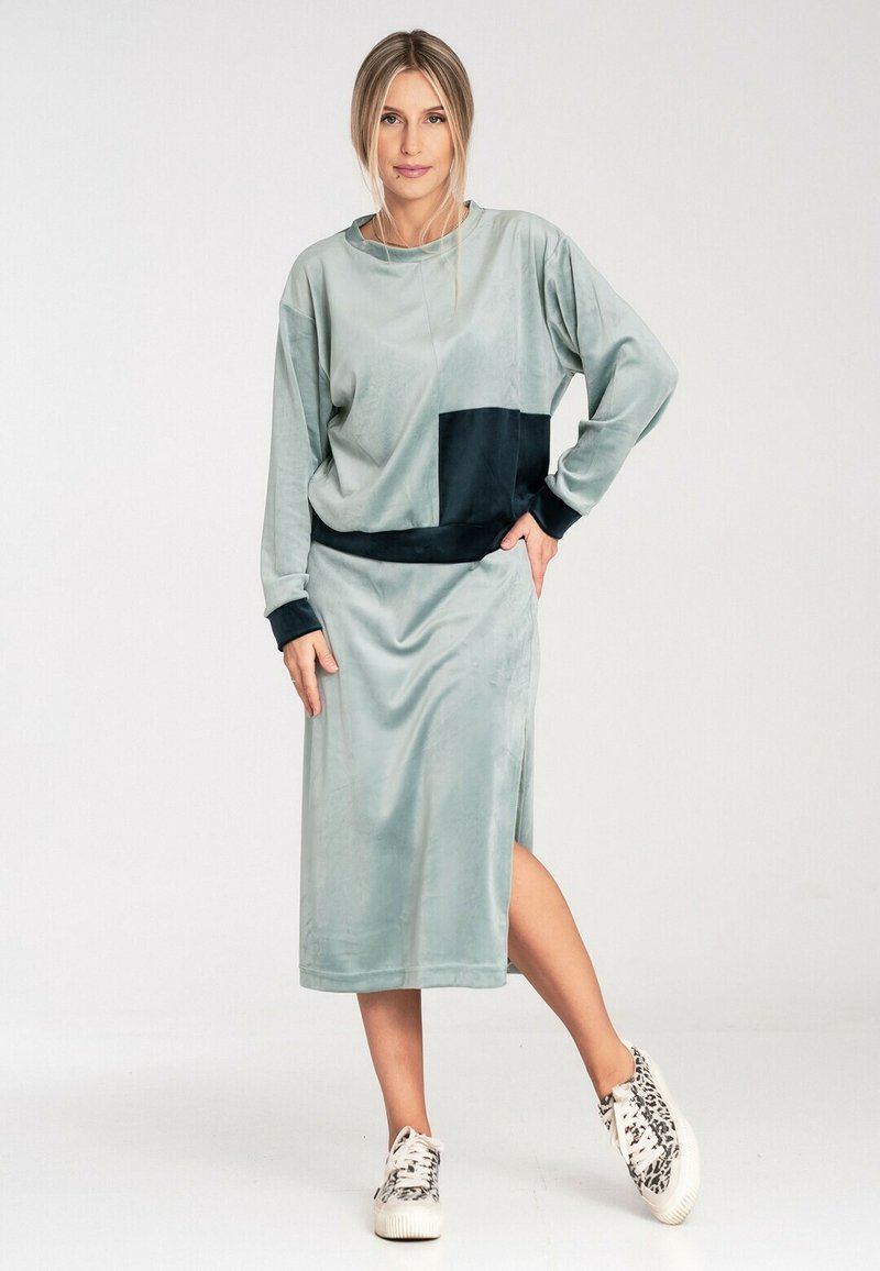 Jasnozielona sukienka z weluru z czarnym panelem, długimi rękawami i bocznym rozcięciem, zestawiona z białymi trampkami. Gładka tekstura i luźny krój.