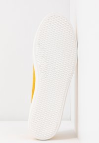 Semelle d'une basket jaune montrant une semelle extérieure en caoutchouc blanche avec motif en chevrons et marquage taille 37, sur fond blanc.