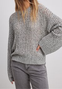 Femme portant un pull gris ample à maille ouverte avec manches longues et un jean gris taille haute, debout avec une main sur la hanche.