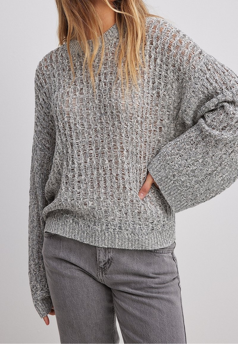 Femme portant un pull gris ample à maille ouverte avec manches longues et un jean gris taille haute, debout avec une main sur la hanche.