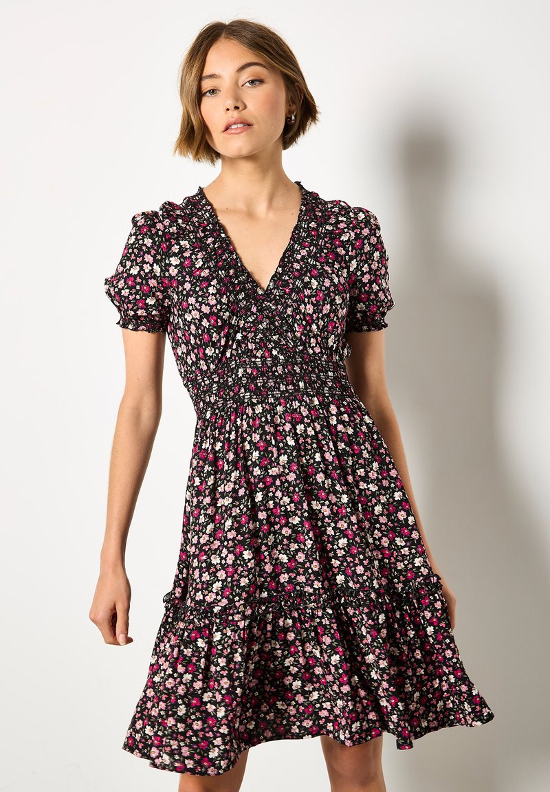 Musta kukallinen mekko, jossa on syvä kaula-aukko, rypytetty yläosa ja puffihihat. Kankaassa on pehmeä tekstuuri, jossa on vaaleanpunaisia ja valkoisia kukkakuvioita.