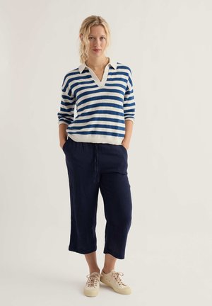 Nő áll kézzel a tengerészkék nadrág zsebében, kék-fehér csíkos pulóvert és bézs színű tornacipőt visel világos háttér előtt.