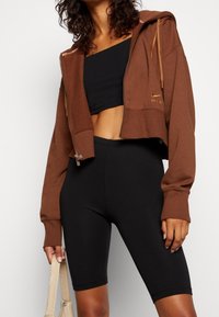 Krótkie brązowe bluza z kapturem na zamek błyskawiczny z złotym logo, zestawiona z czarnymi dopasowanymi szortami i beżową torebką. Gładka tekstura materiału.
