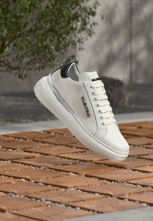 Baskets en cuir blanc avec accents noirs, présentant un bout rond, un laçage frontal, et un logo sur le côté. Texture lisse et design minimaliste.