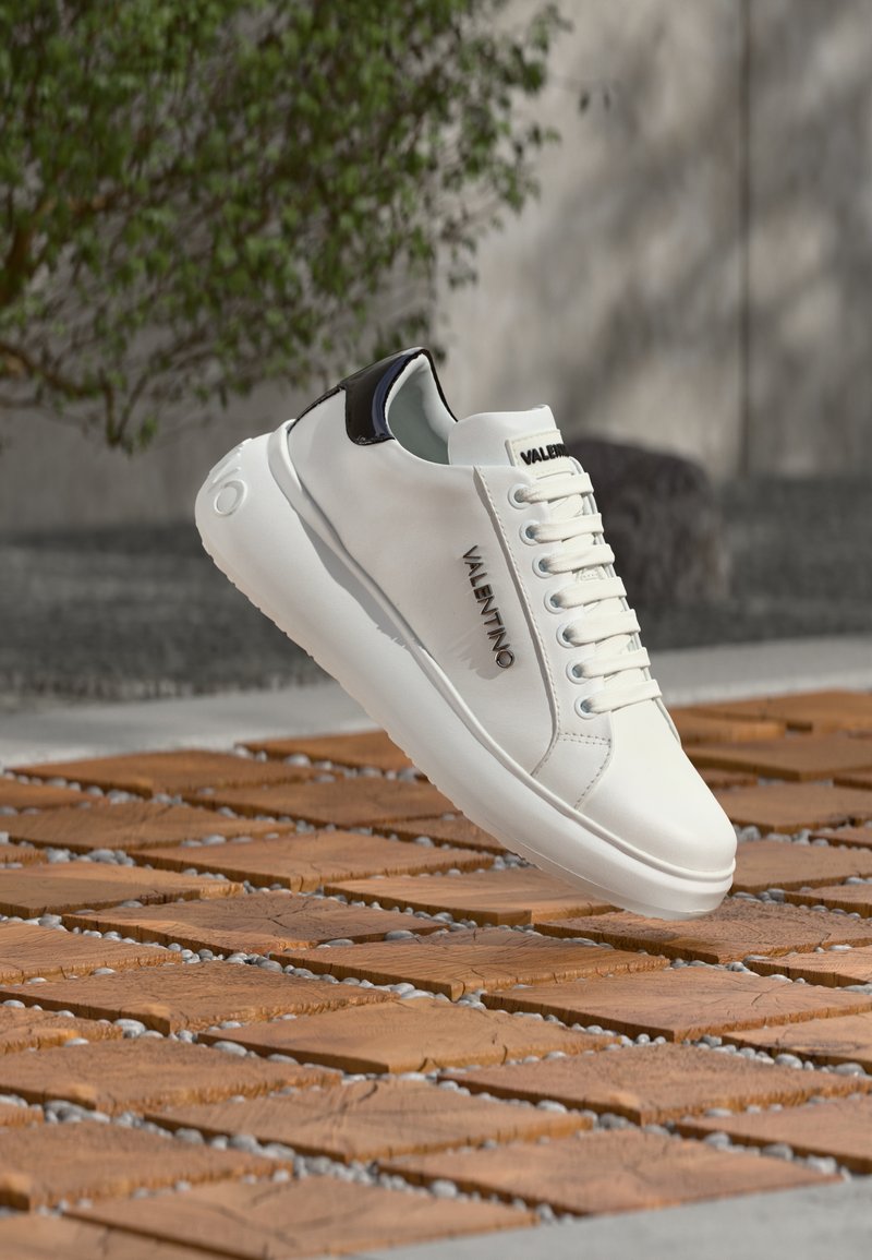 Baskets en cuir blanc avec accents noirs, présentant un bout rond, un laçage frontal, et un logo sur le côté. Texture lisse et design minimaliste.