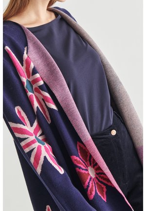 Châle en laine bleu marine avec des motifs floraux roses et blancs ; bordure rose avec des détails texturés. Superposé sur un haut et un pantalon sombres.