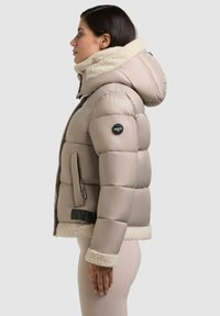 Beige Daunenjacke mit einem flauschigen cremefarbenen Kragen und Manschetten, seitlichen Taschen, einem Logoschild und einem Reißverschluss auf der Vorderseite.
