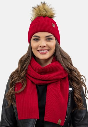 Cappello di maglia rosso con pompon in pelliccia sintetica e una sciarpa spessa abbinata. Entrambi presentano una texture a costine e un piccolo etichetta con logo sulla sciarpa.