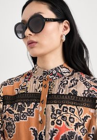 Ochelari de soare rotunzi, negri, cu o ramă groasă, purtați cu o bluză cu model, ce prezintă motive florale, accente din dantelă și nasturi din lemn.