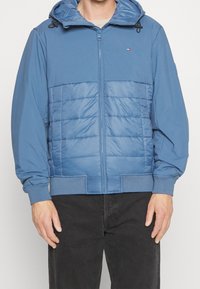 Muž oblečený v modré zipové mikině s kapucí a vycpaným předním dílem, s žebrovanými manžetami, v kombinaci s černými kalhotami, stojící na jednobarevném pozadí.