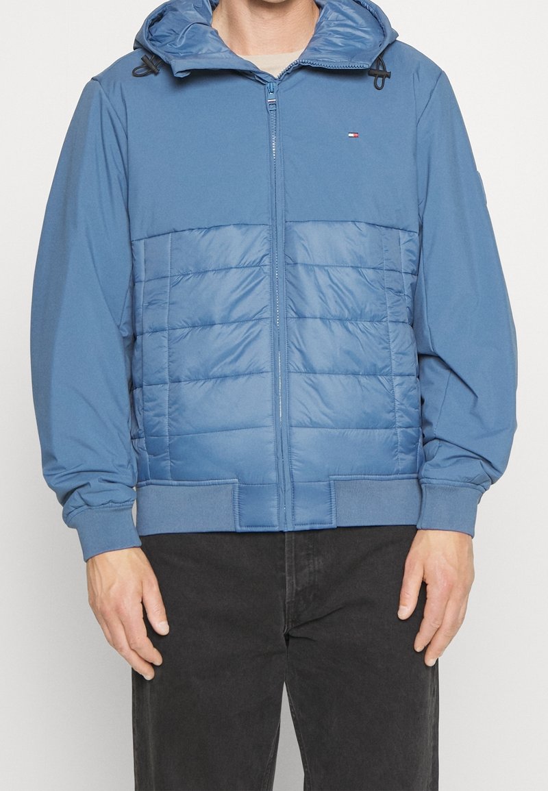 Muž oblečený v modré zipové mikině s kapucí a vycpaným předním dílem, s žebrovanými manžetami, v kombinaci s černými kalhotami, stojící na jednobarevném pozadí.