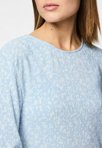 Top azul claro con pliegues verticales y pequeños patrones florales blancos. Presenta un escote redondo y una textura suave y lisa.