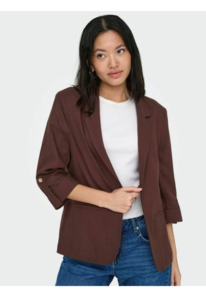 Ung kvinde med langt mørkt hår iført en brun blazer over en hvid top og blå jeans, poserer med den ene hånd, der holder blazeren.