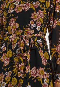 Blumenkleid mit schwarzem Hintergrund, kunstvollen rosa und gelben Blumen, tailliertem Schnitt und passendem Stoffgürtel. Lange, durchsichtige Ärmel.