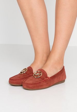 Mocasines de ante en rojo óxido con un logo circular dorado en la parte delantera, con suela plana y detalles cosidos.