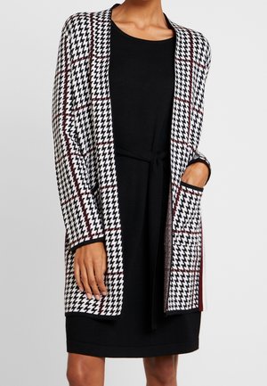 Kvinde iført en sort knælængde kjole og en sort, hvid og rød hundetandsmønstret cardigan med åbent front og lommer.