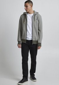 Grijze zip-up hoodie met een gestructureerd patroon, groene accenten en gele koorden, gecombineerd met zwarte broek en zwarte sneakers.