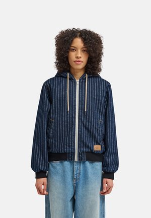 Jeune personne aux cheveux bouclés portant un sweat à capuche zippé rayé bleu avec cordons beige et un jean bleu clair ample, debout devant un fond uni.