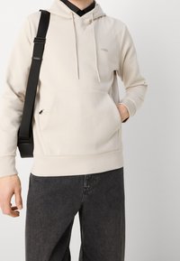 Felpa con cappuccio beige, dotata di tasca a marsupio, polsini a costine e un piccolo logo sul petto. Indossata con jeans grigio scuro e una tracolla nera.