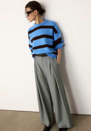 Blue and brown striped oversized sweater paired with tailored gray pleated trousers. Model wears dark sunglasses and a black belt.