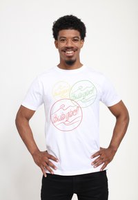 Vit bomull t-shirt med tre överlappande cirkeldesigner i röd, grön och gul, var och en med "Babybel"-text och vågiga accenter.