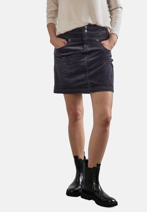 Frau trägt einen schwarzen Cord-Minirock, beige langärmliges Oberteil, Hände in den Taschen, und schwarze glänzende Leder-Stiefeletten.