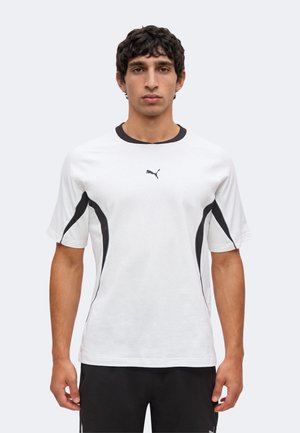 Άνδρας που φοράει λευκό T-shirt Puma με μαύρες πλευρικές πινελιές και γιακά, στέκεται μπροστά σε απλό φωτεινό φόντο.