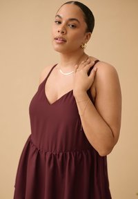 Femme aux cheveux bruns attachés, portant une robe bordeaux sans manches et des bijoux en or, touchant son épaule sur un fond beige.