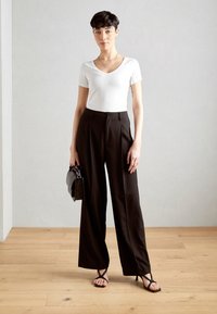 T-shirt blanc ajusté à col V associé à un pantalon large noir taille haute. Le modèle tient un sac à main noir texturé et porte des sandales noires à lanières.