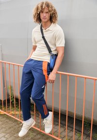 Maglietta polo beige a costine, pantaloni della tuta navy con pannelli laterali arancioni e sneakers bianche con tre strisce, in piedi vicino a una barriera arancione.