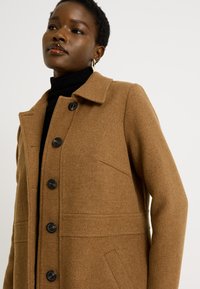 Abrigo de mezcla de lana marrón con cuello tipo solapa, detalles de costura vertical, cuatro botones negros y bolsillos laterales. Tejido texturizado.