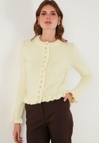 Vrouw in een crèmekleurige cardigan met ruches en knopen, met donkerbruine broek, staand tegen een effen lichte muur.