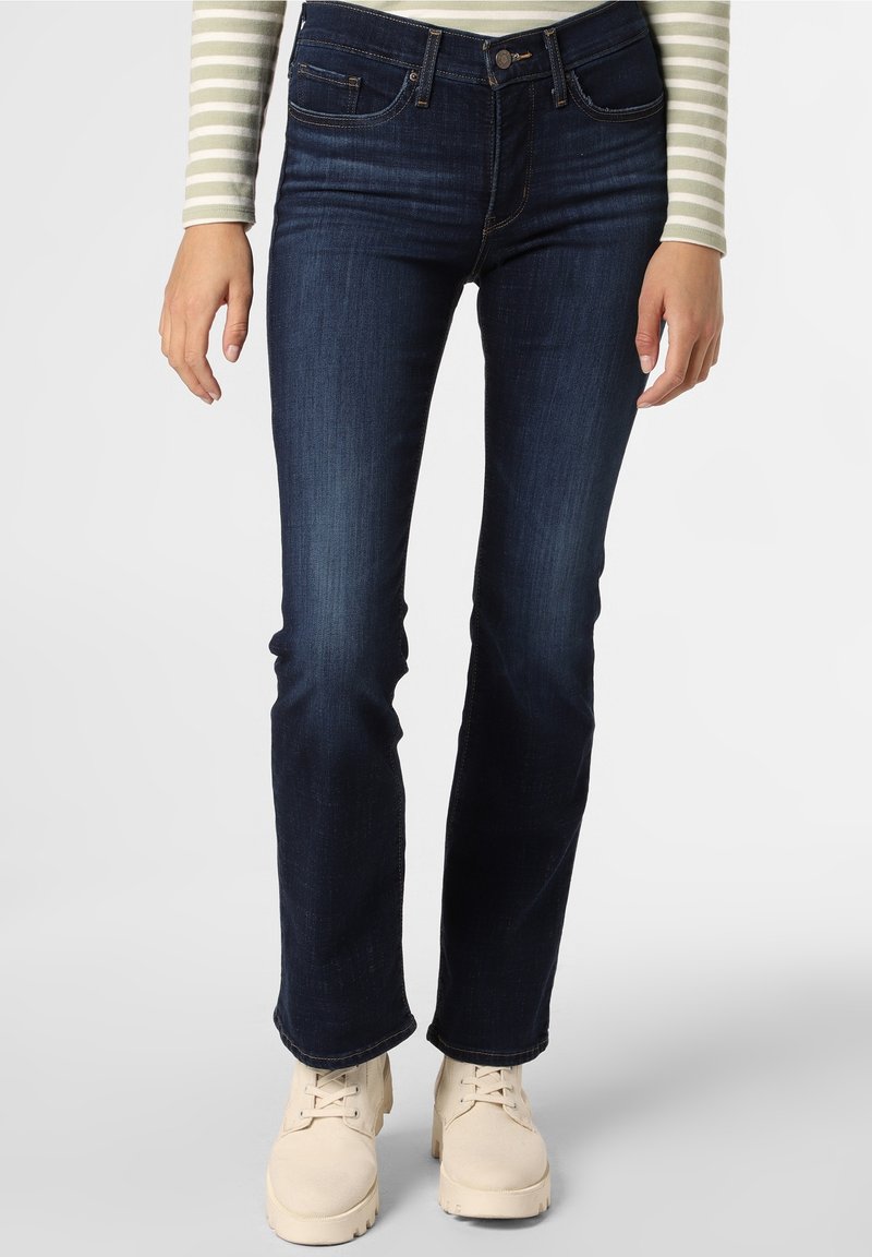 Jean en denim bleu foncé avec une coupe droite, cinq poches et une taille mi-haute, associé à des bottes beige clair.