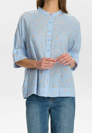 Osoba nosząca jasnoniebieską koszulę na guziki z małymi różowymi kwiatowymi wzorami i podwiniętymi rękawami, zestawioną z niebieskimi dżinsami.