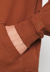 Felpa marrone con polsini a coste e una tasca laterale. Include un piccolo logo ricamato sul polsino in una tonalità abbinata. Morbida al tatto.