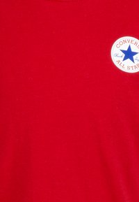 Maglietta rossa di cotone con scollo rotondo, caratterizzata da un logo circolare in blu e bianco con un motivo a stella e la scritta "Converse All Star".