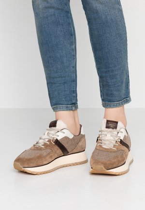 Personne portant des baskets beige et marron avec des détails blancs et en fourrure, associées à un jean en denim bleu, debout sur un sol clair.