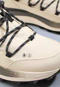 Beżowe buty trekkingowe z teksturowaną tkaniną, czarnymi sznurowadłami oraz symbolami wskazującymi na oddychalność i wodoodporność. Gumowa podeszwa zewnętrzna zapewniająca przyczepność.