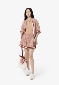 Ung kvinna som bär mönstrad öppen skjorta, matchande shorts, beige pikétröja, vita sneakers och håller en vit tygkasse med matchande scarf.