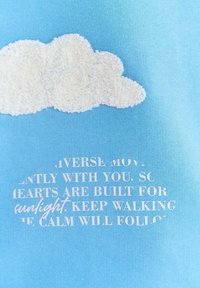 Jasnoniebieska tkanina z wzorem puszystej białej chmury i teksturowanymi detalami. Poniżej chmury znajduje się biały tekst z inspirującym przesłaniem.