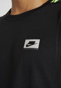 Lähikuva mustasta urheilupaidasta, jossa on pieni valkoinen neliölogo, jossa on musta swoosh ja geometrinen muotoilu rinnassa.