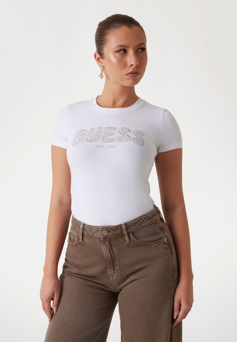 T-shirt bianco a maniche corte con logo "GUESS" in strass argentati e "EST. 1981" sotto, abbinato a jeans marroni a vita alta.